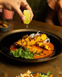 A hand squeezes lemon over tandoori chicken on a sauce and garnished with cilantro.